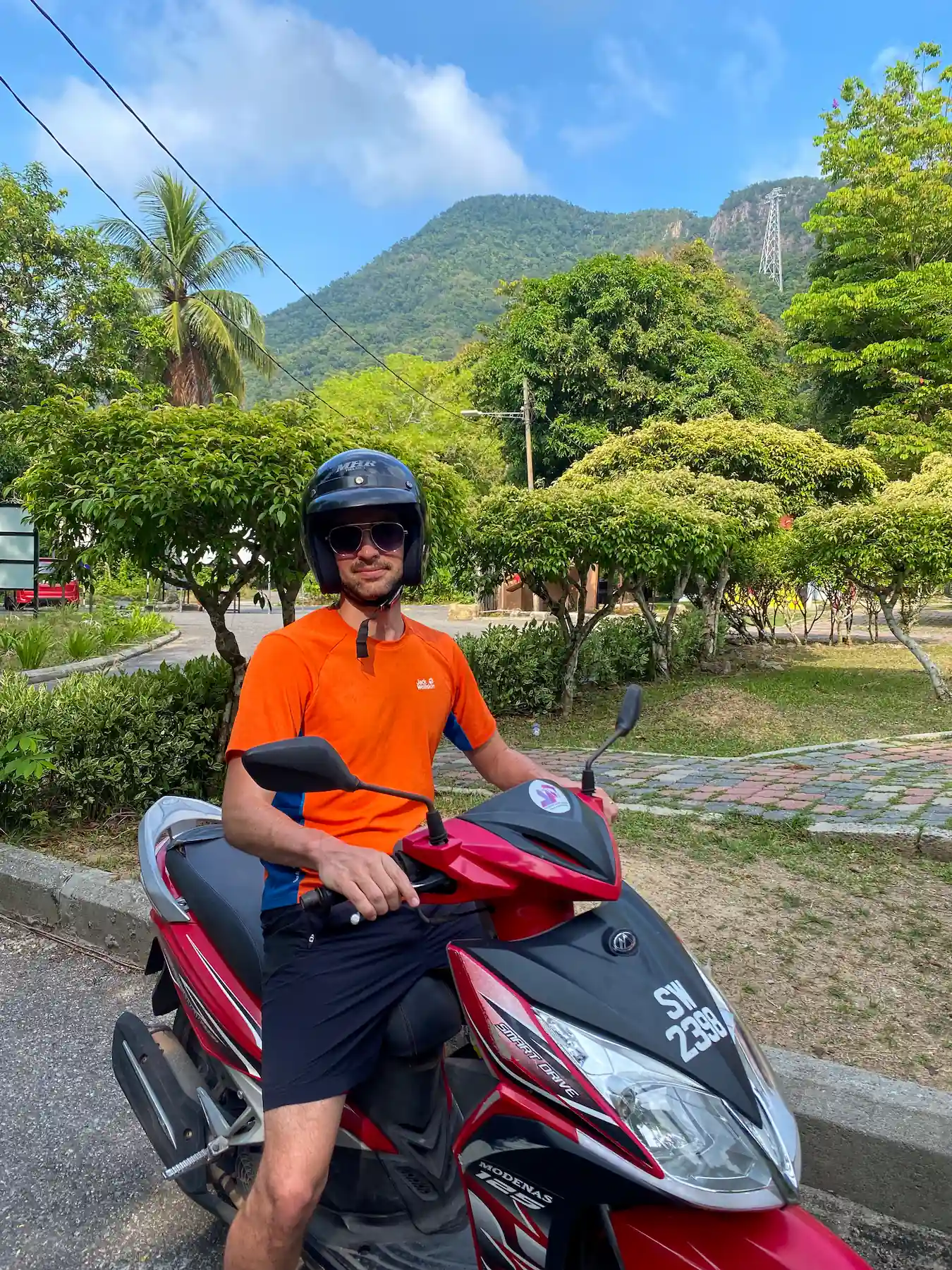 Man in orange shirt sitting on red moped with tropical mountain landscape in background